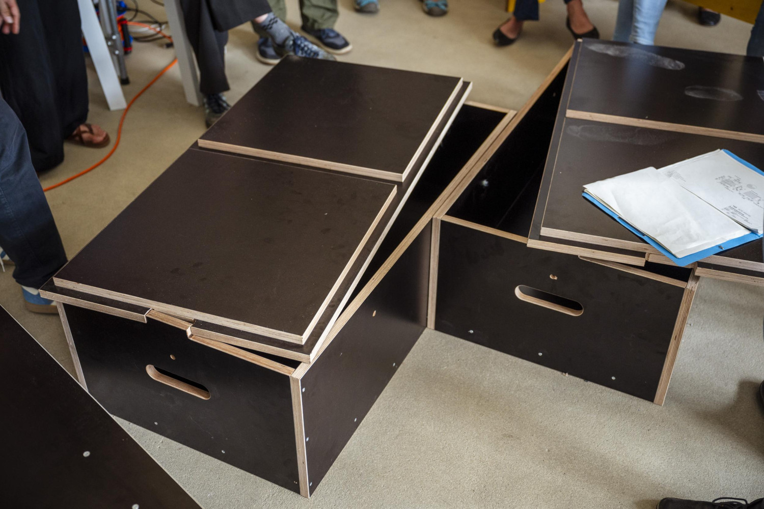 Schwarze Kisten mit Holzrand, teils offen, auf einem hellen Boden, umgeben von Menschen.