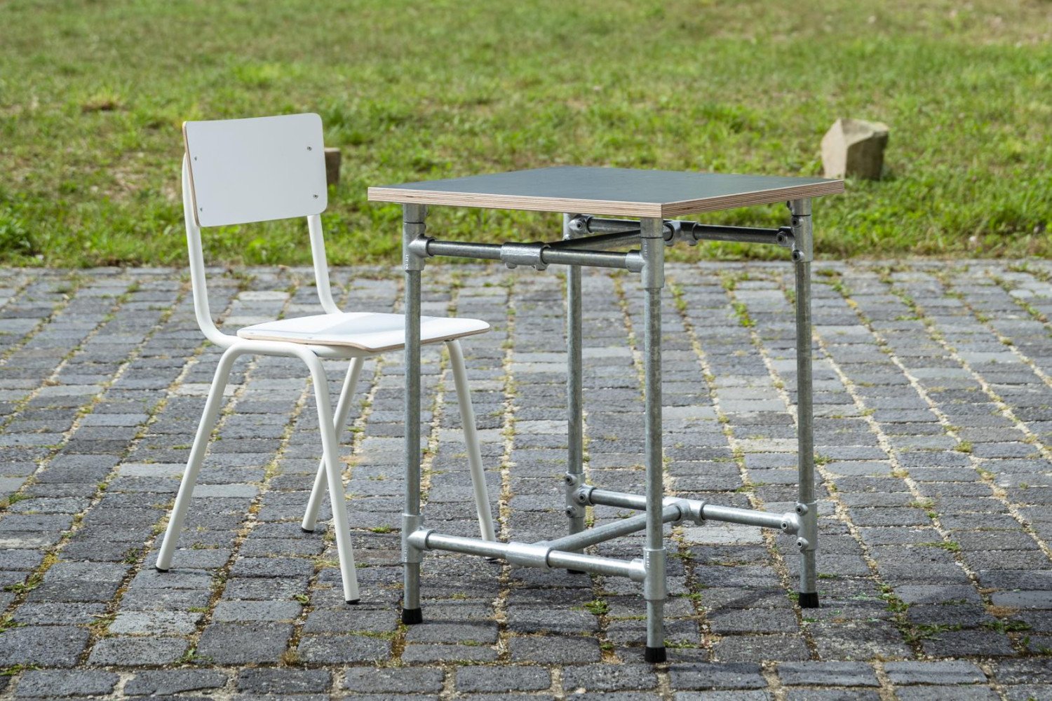 Ein Tisch und ein Stuhl stehen auf einem Pflasterweg, umgeben von grünem Umland und bewölktem Himmel.