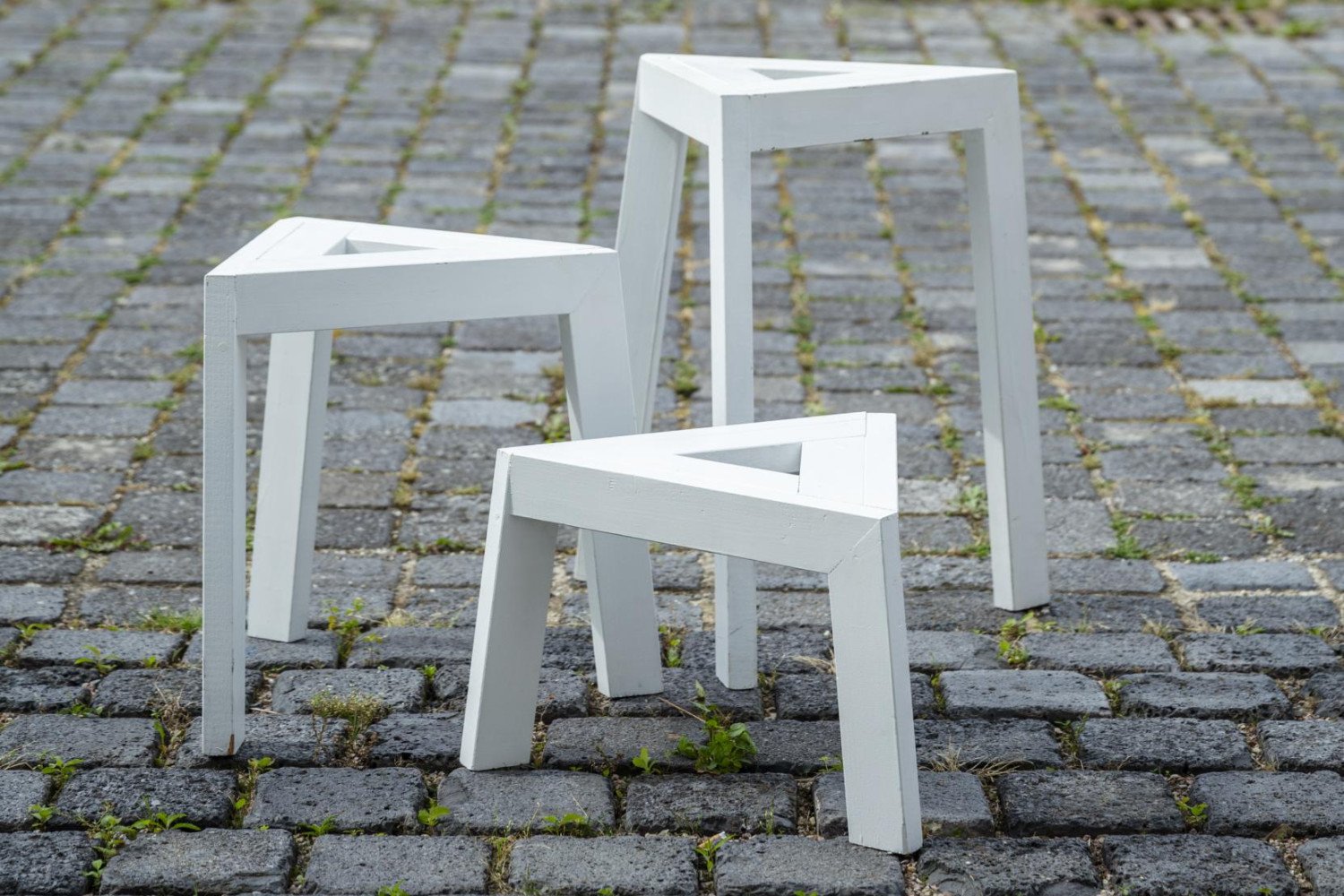 Drei moderne, weiße Hocker stehen auf einem Pflastersteinboden in einer grünen Wiese.