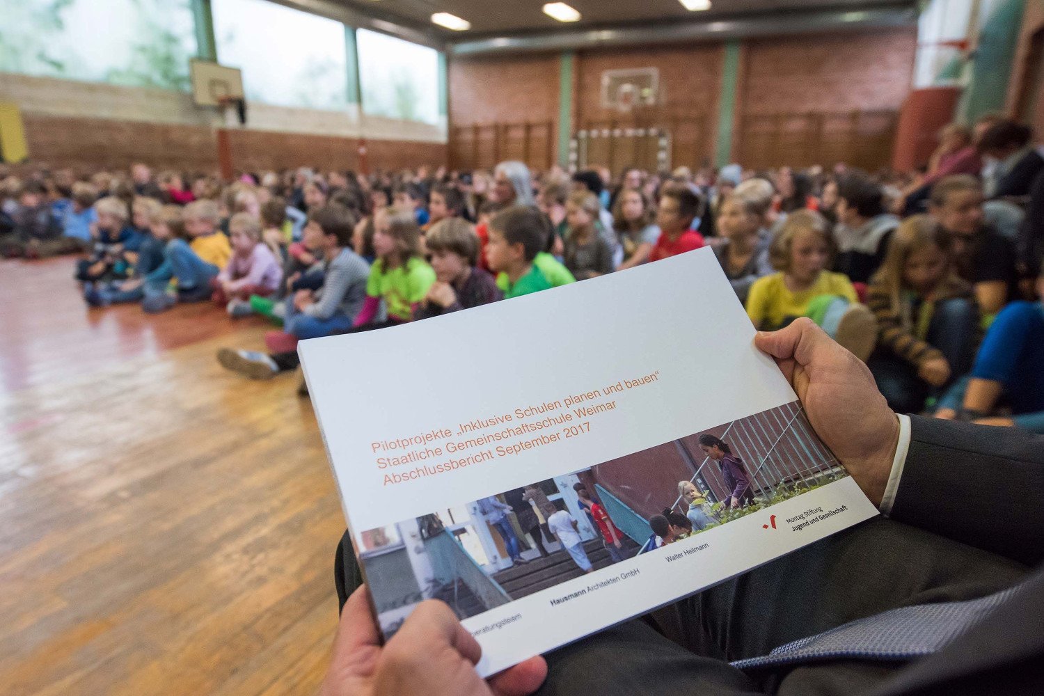 Kinder Schule sitzen bei einer Veranstaltung in der Turnhalle. Die druckfrische Broschüre des Phase Null Berichtes wird in das Bild gehalten.